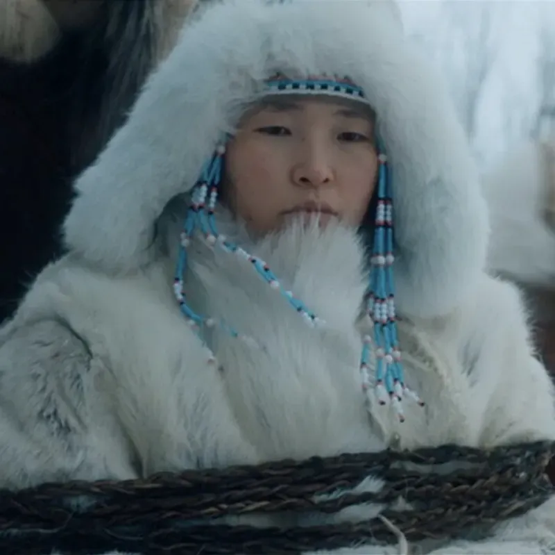 Film still: a child in a white fur-trimmed hood with beaded ornaments in a snowy landscape